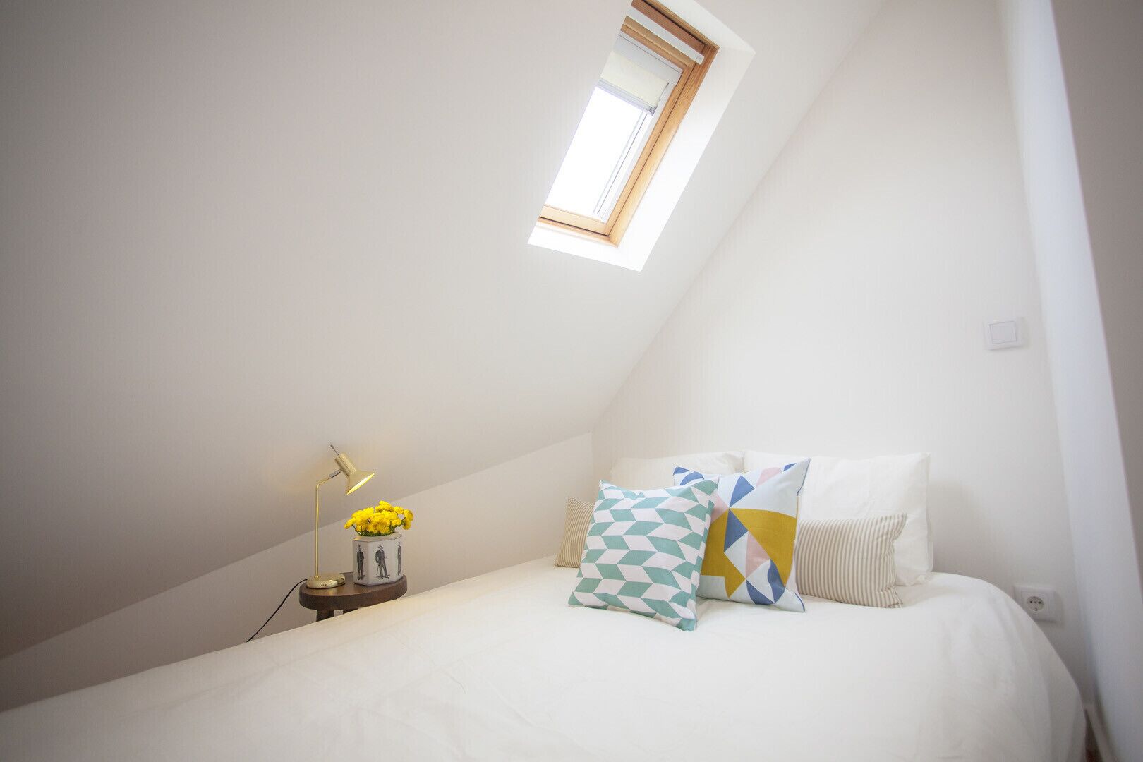 A bedroom with a sloped ceiling, a skylight, a bedside table with a lamp, and a bed with patterned pillows.