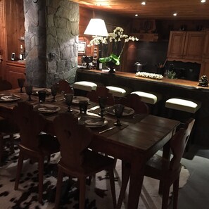 Un coin repas avec un mur en pierre, des chaises en bois, une table dressée pour quatre personnes et une cuisine en arrière-plan.