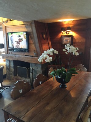 A wooden dining table with a vase of white orchids, a fireplace, and a television mounted on the wall.