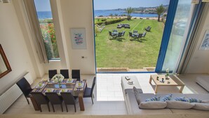A spacious living area with a dining table set for four, a sofa, and a view of a well-maintained lawn and water body. - Protaras Fig Tree Bay Seafront Villa Infinite Aretousa (Protaras)