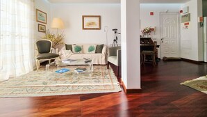 Ein Wohnzimmer mit einer Couch, Sesseln, einem Couchtisch und einem Teppich. Eine Tür führt in einen anderen Raum, und auf einem Beistelltisch steht eine Lampe.