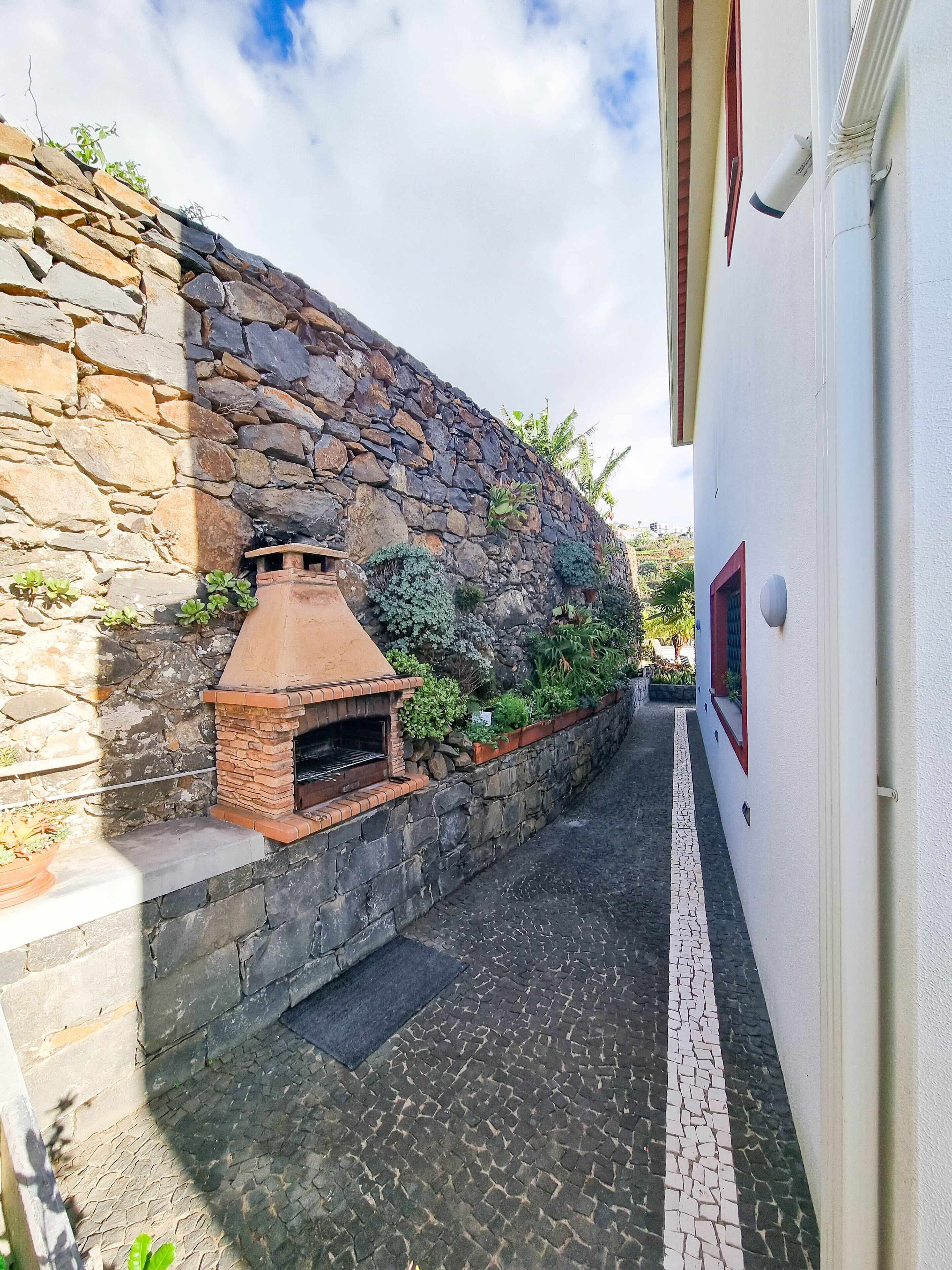 A narrow alley with a stone wall on one side and a white building on the other. A brick oven is built into the stone wall.