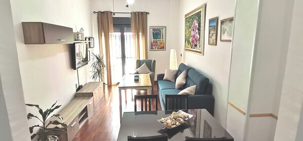 A living room with a sofa, a dining table, and a coffee table. There is a plant on the left and a lamp on the right.