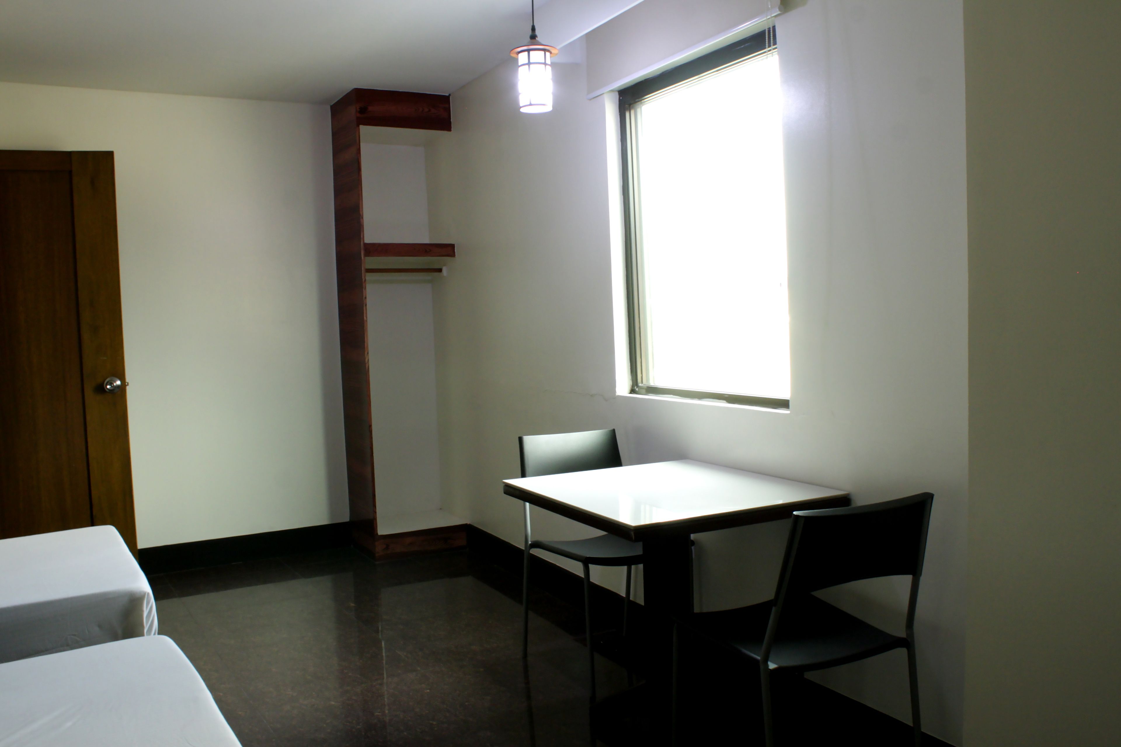 A small, sparsely furnished room with a single table and two chairs, a window with natural light, and a door.