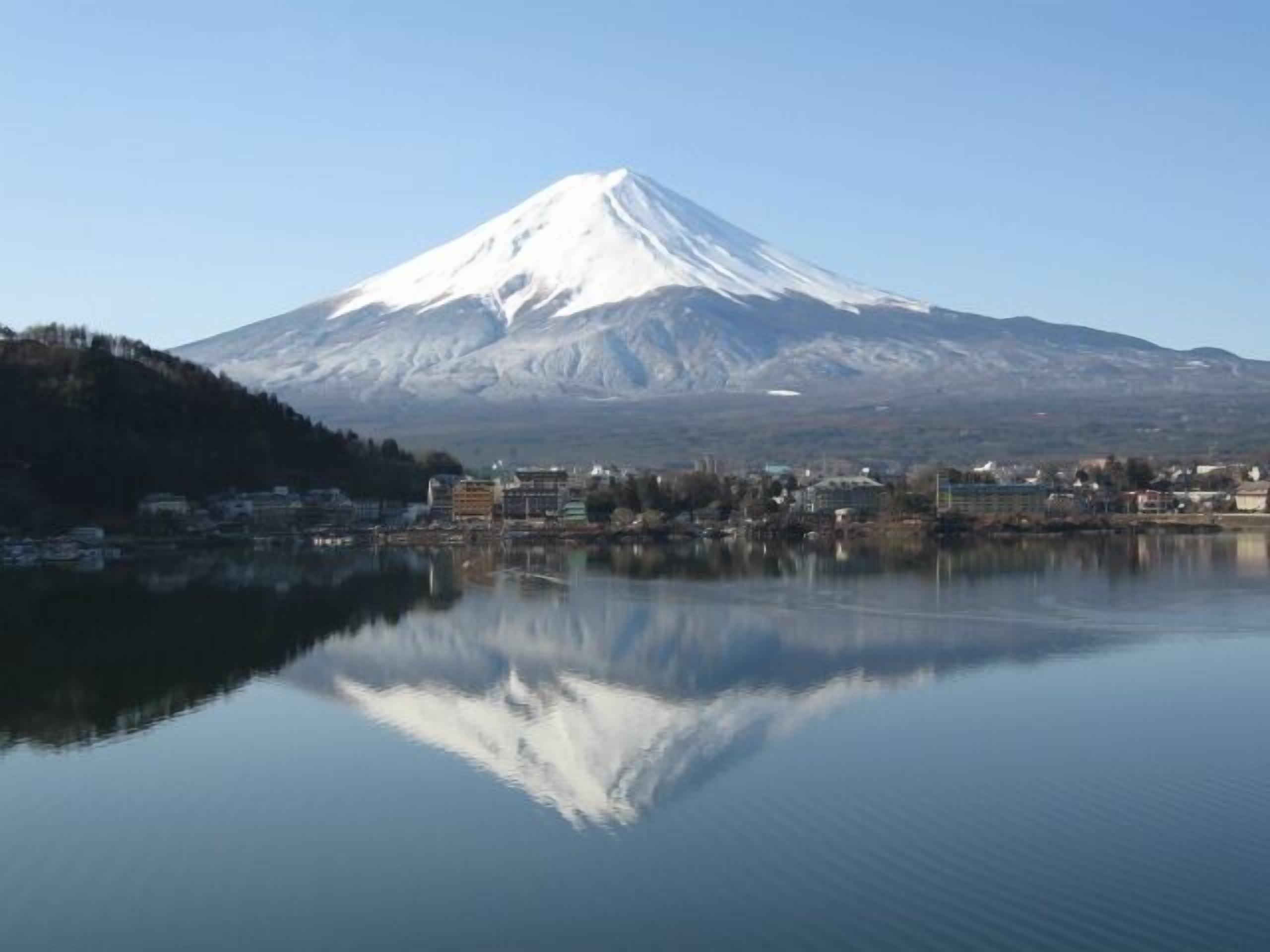 Montanha coberta de neve com reflexo nítido em águas calmas, cercada por edifícios.