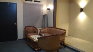 Una habitación con una puerta oscura, una ventana cerrada con persianas, dos sillones de cuero, una mesita con una botella y una lámpara fijada a la pared.