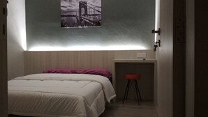 Una habitación pequeña y tenuemente iluminada con una cama, un taburete y una estantería en la pared.