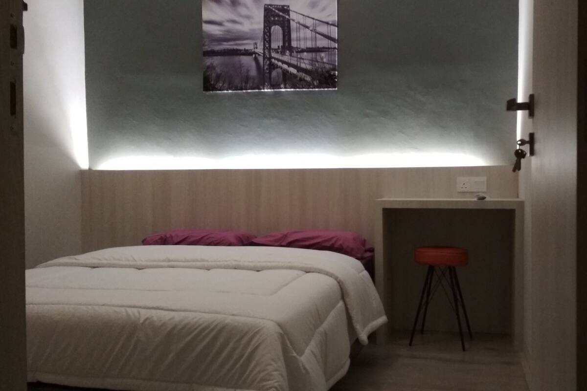 A small, dimly lit room with a bed, a small stool, and a wall-mounted shelf.