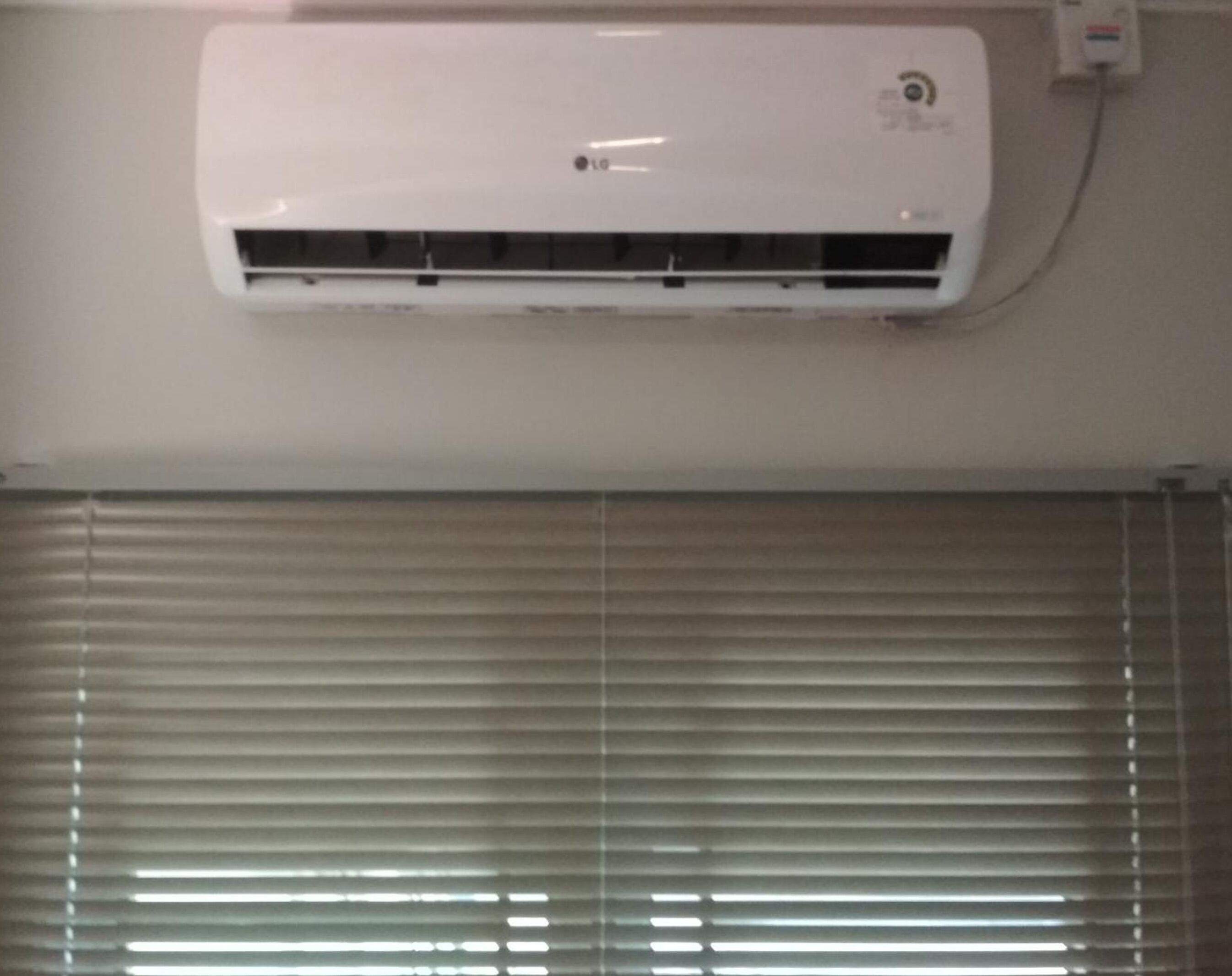 A white air conditioning unit mounted on a wall above a window with horizontal blinds.