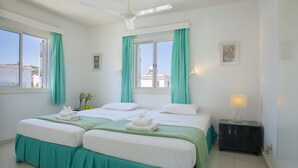 Habitación de hotel con dos camas, cortinas color turquesa, un ventilador de techo, una mesita de noche con una lámpara y una ventana con vistas a edificios.