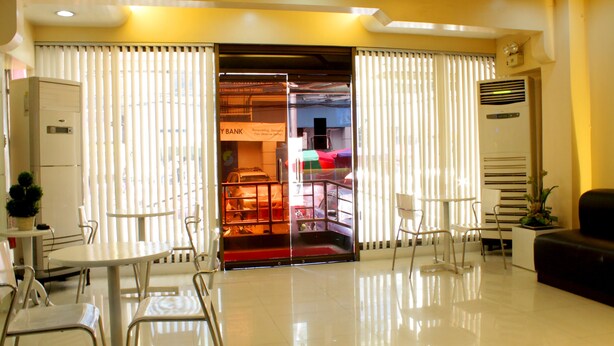 A hotel lobby with a reception desk, a seating area with chairs and a table, and a large window with blinds. - Vista Apartelle & Restaurant (Makati)