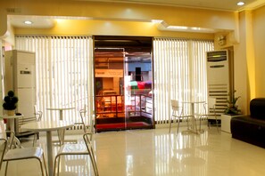 A hotel lobby with a reception desk, a seating area with chairs and a table, and a large window with blinds.