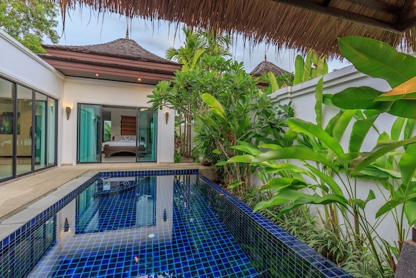 A modern pool with blue tiles, surrounded by lush greenery and a white building with large windows.
