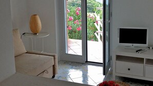 Habitación con puerta de vidrio corrediza que da a un balcón con vistas a plantas y flores. Hay un sofá blanco, una mesita con lámpara y un televisor sobre un mueble.
