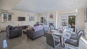 A modern living room with a dining area, a fireplace, and large windows. - Protaras Villa Theodora 1 - CHG (Paralimni)