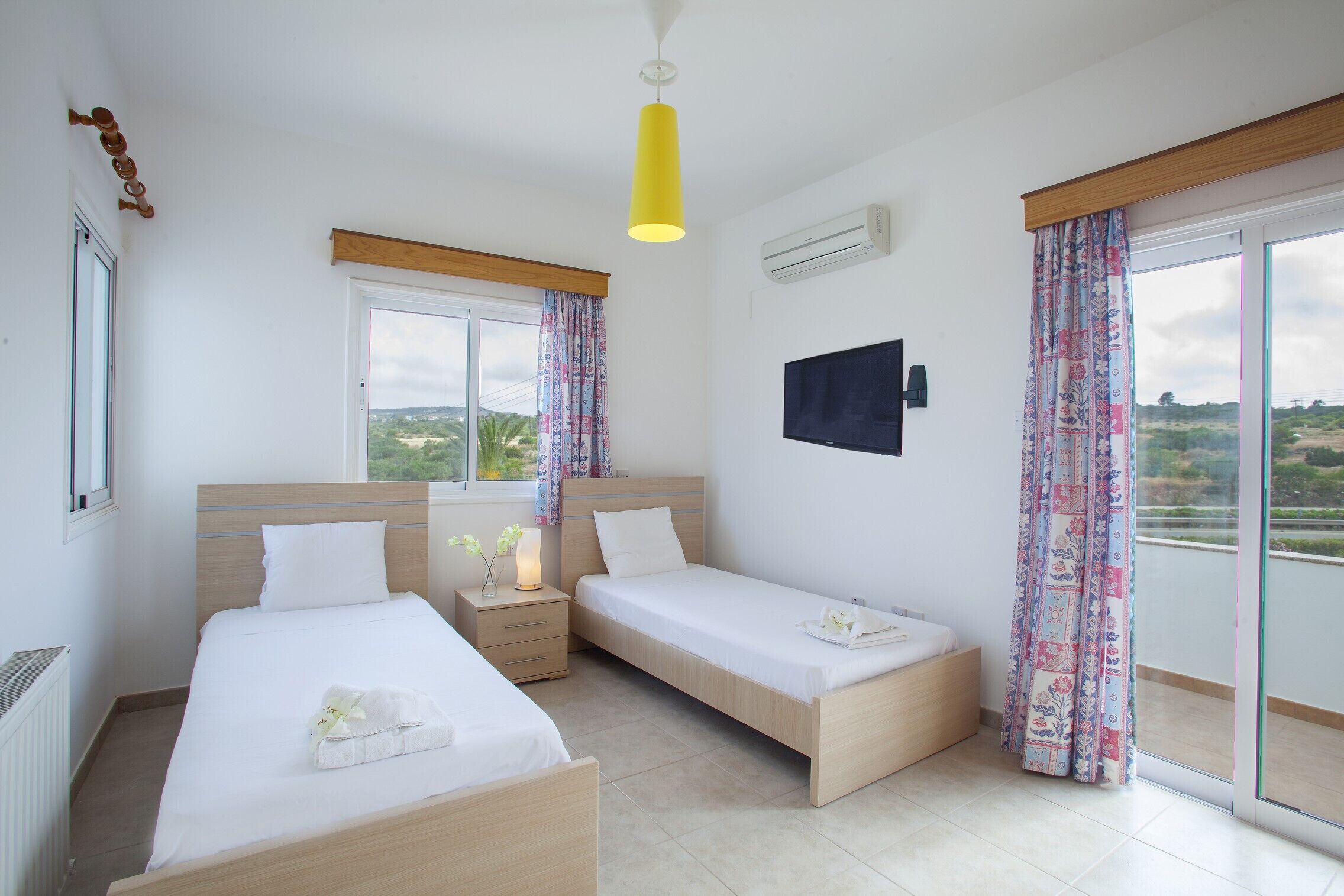 A hotel room with two beds, a TV, a window with curtains, and a view of the outdoors.