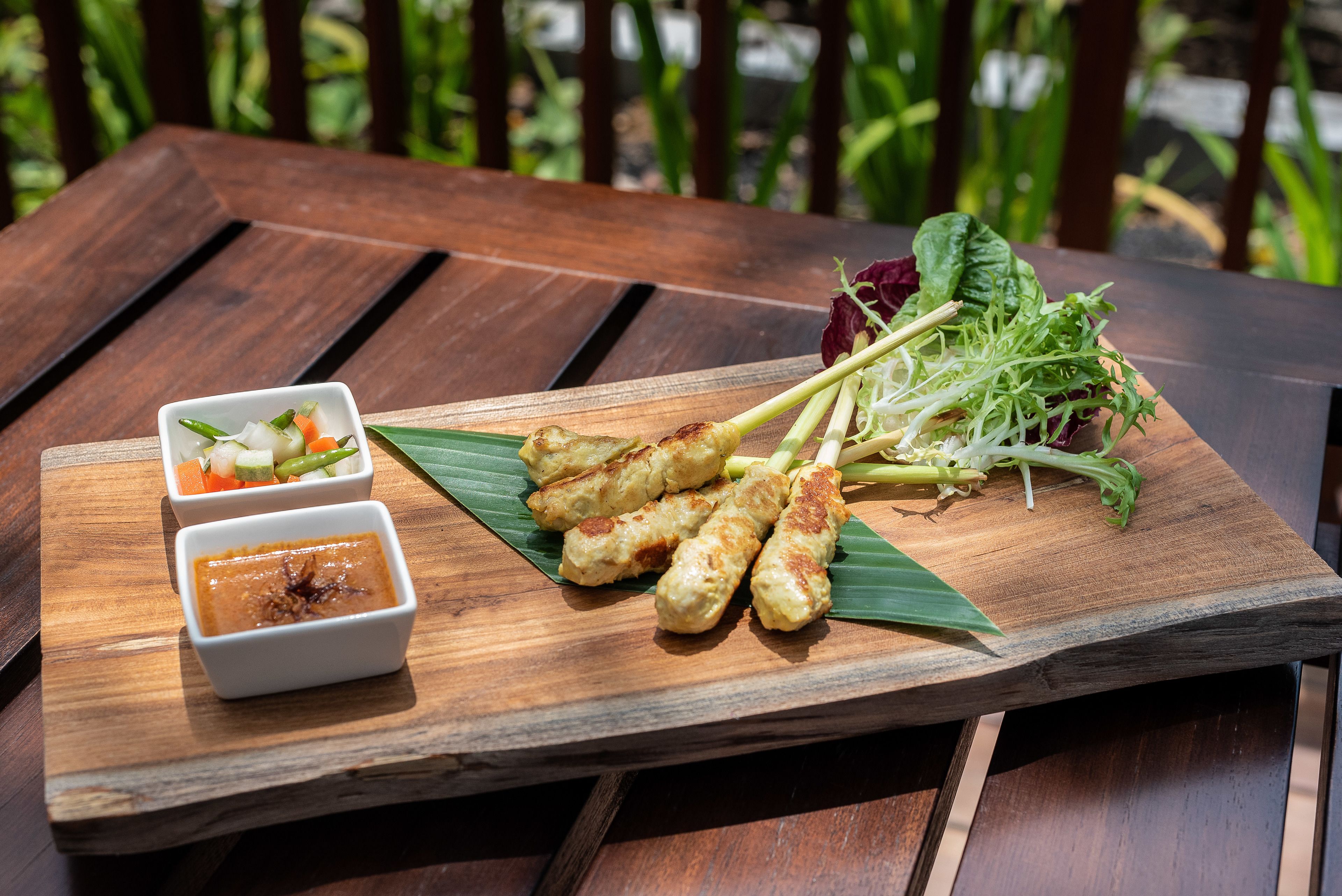 A wooden table with grilled meat skewers, fresh vegetables, and two small bowls of dipping sauces.