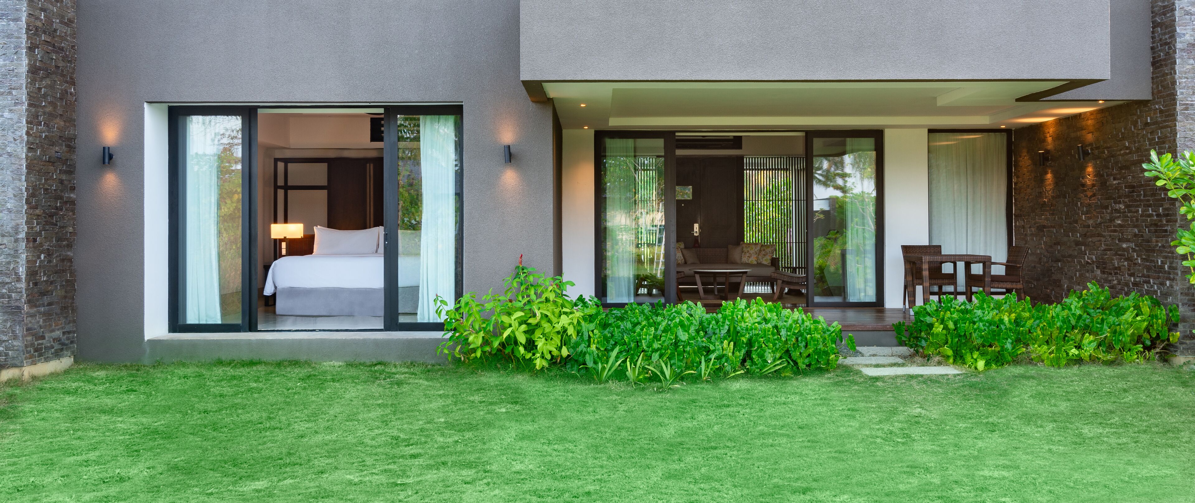 A modern hotel room with a large sliding glass door leading to a balcony, a dining area with a table and chairs, and a well-maintained garden.