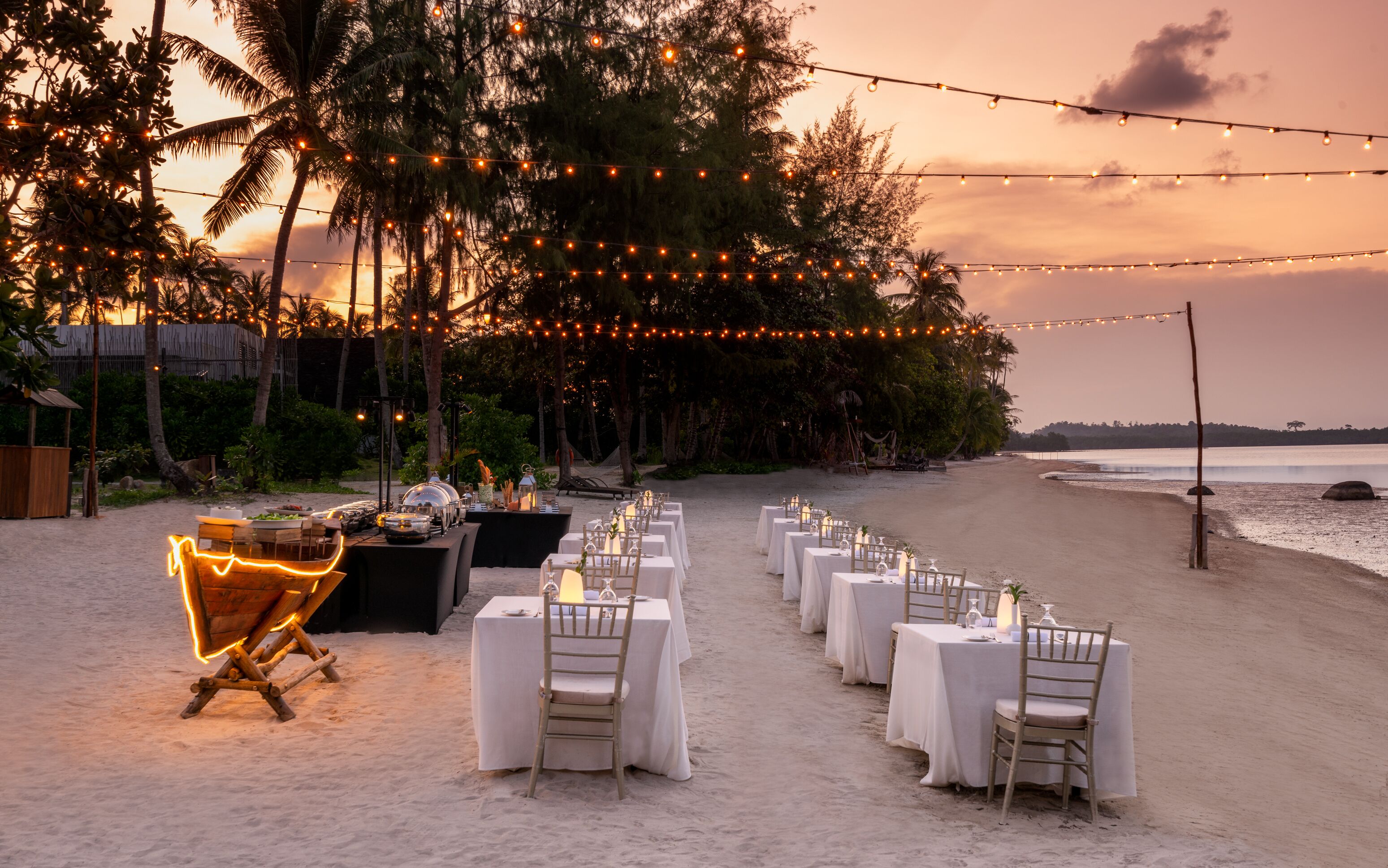 A beachside dining setup with tables and chairs, string lights, and a view of the ocean.