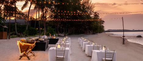 A beachside dining setup with tables and chairs, string lights, and a view of the ocean.