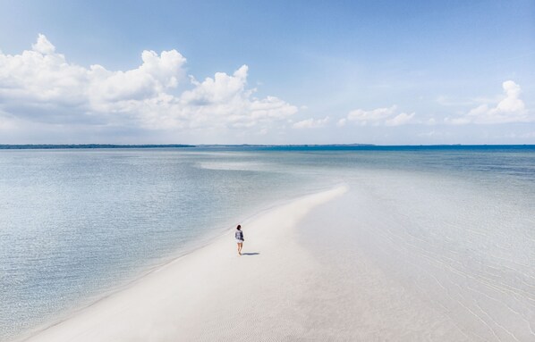 A person walking on a wide, sandy beach with clear blue water and a distant shoreline. - The Residence Bintan (Bintan)