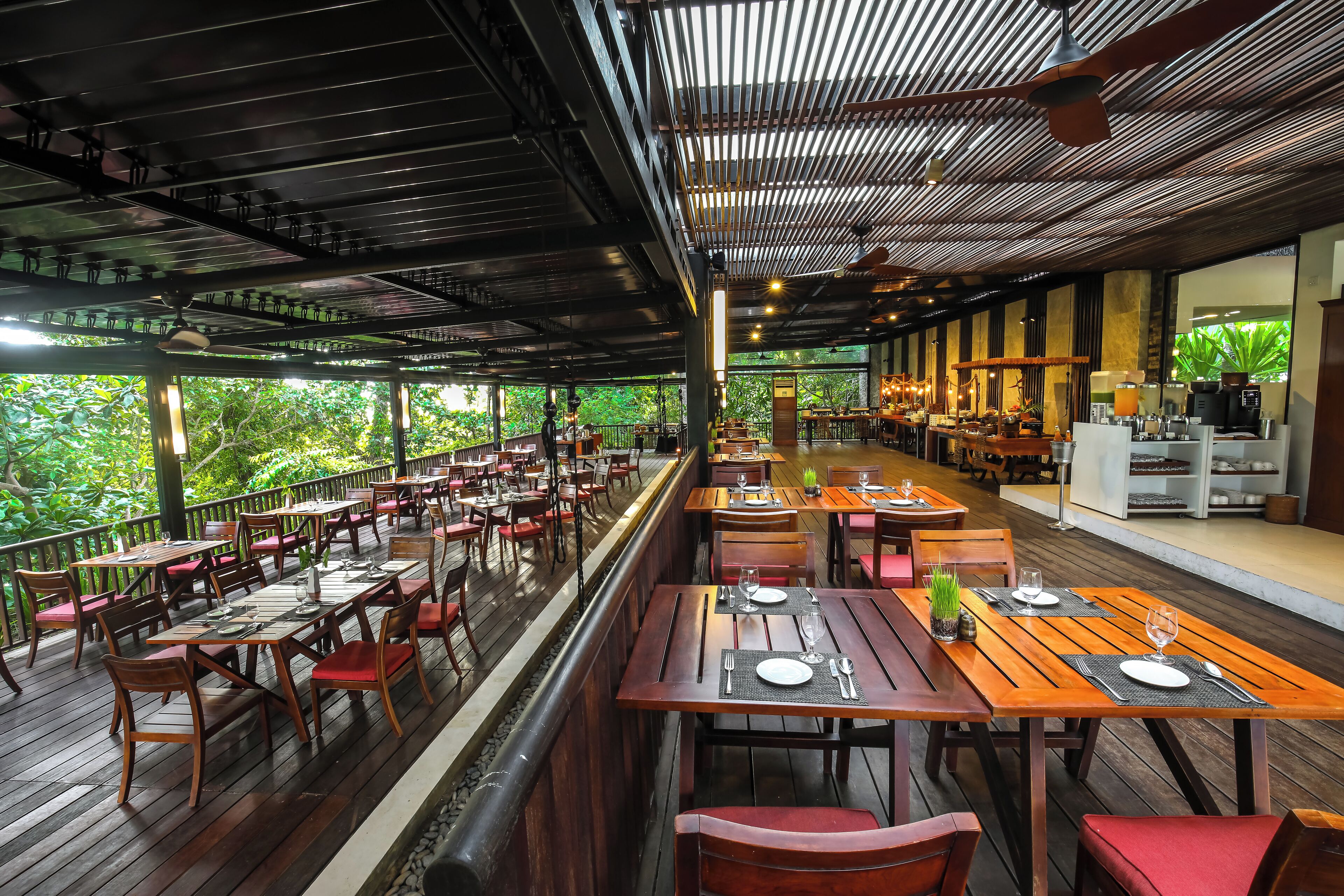 An indoor restaurant with wooden tables and chairs, a high ceiling with exposed beams, and large windows overlooking greenery.