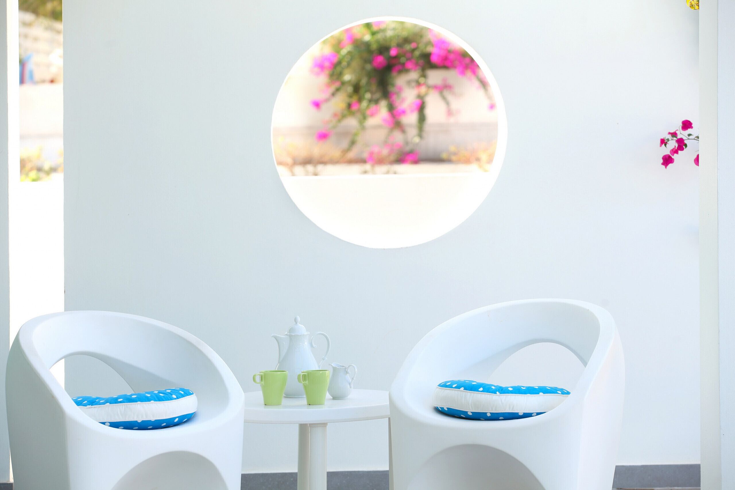 Ein Zimmer mit zwei weißen Stühlen, einem kleinen Tisch mit einer weißen Karaffe und zwei grünen Tassen sowie einem runden Fenster mit unscharfem Blick auf rosa Blumen.