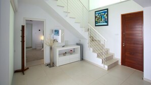 Un hall amplio con una escalera que sube a un piso superior, una consola blanca con adornos y una puerta de madera.