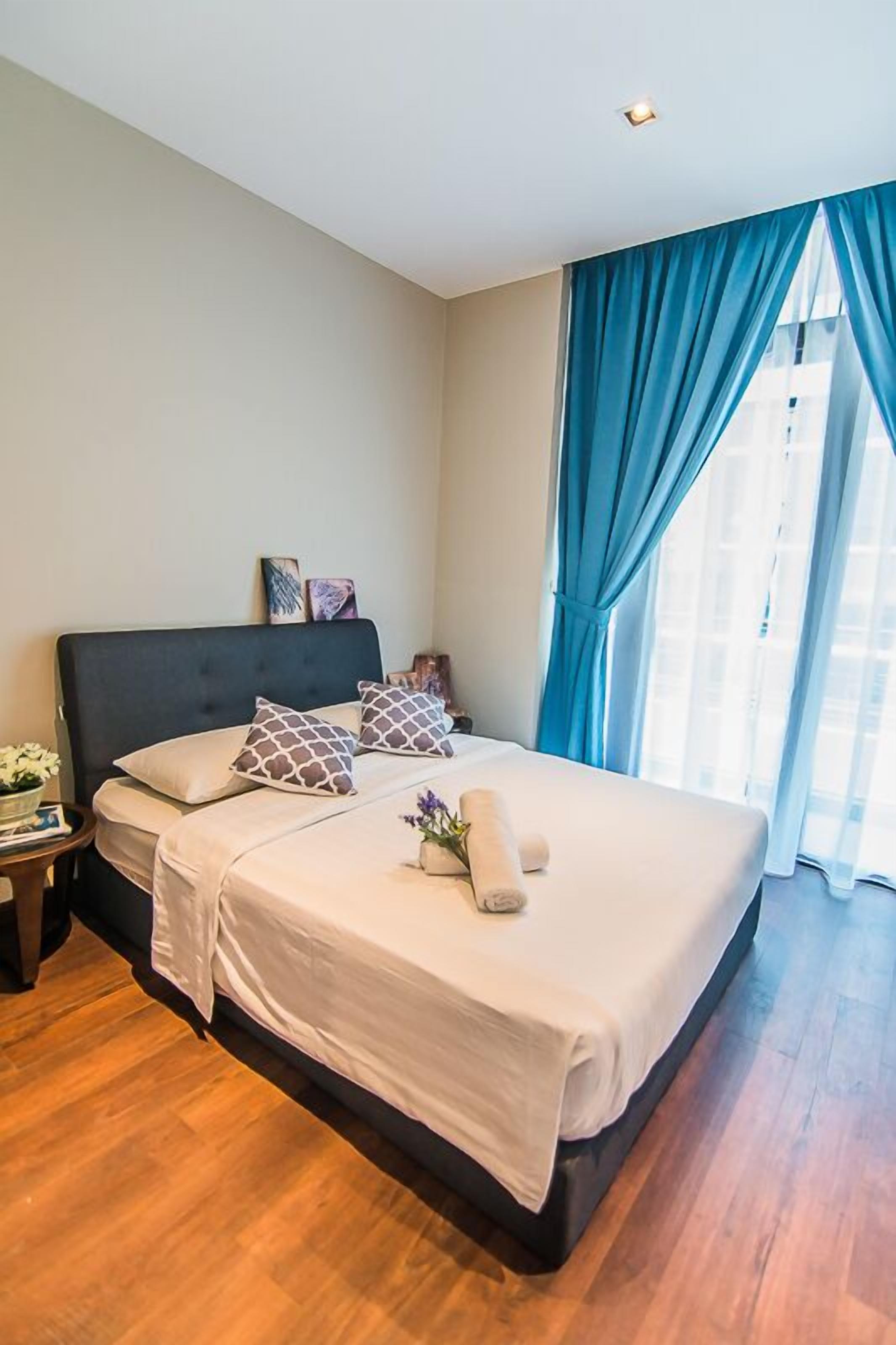A bedroom with a large bed, a nightstand, a vase with flowers, and a window with blue curtains.