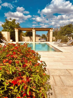 Une piscine agrémentée d’une pergola et de chaises longues, entourée de verdure et surplombée par un ciel dégagé.