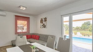 A modern living room with a grey sofa, red cushions, a dining table with a plant, and a view of a swimming pool through a sliding glass door. - Noelene - Cozy house, Private Pool, BBQ, Quiet area, Privacy (Valtura)