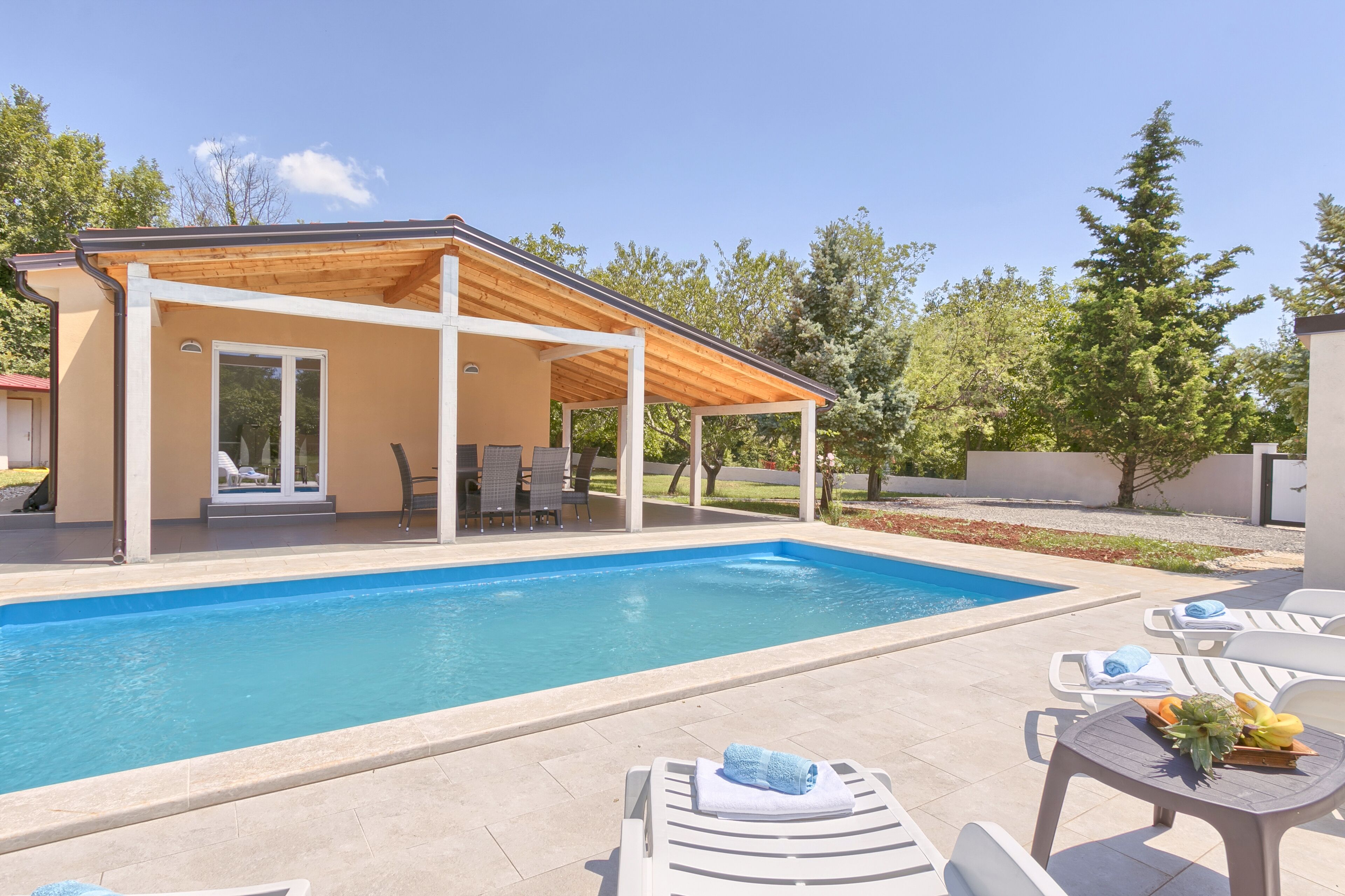 Una piscina con un porche cubierto, sillas de descanso y una mesa con frutas y toallas.