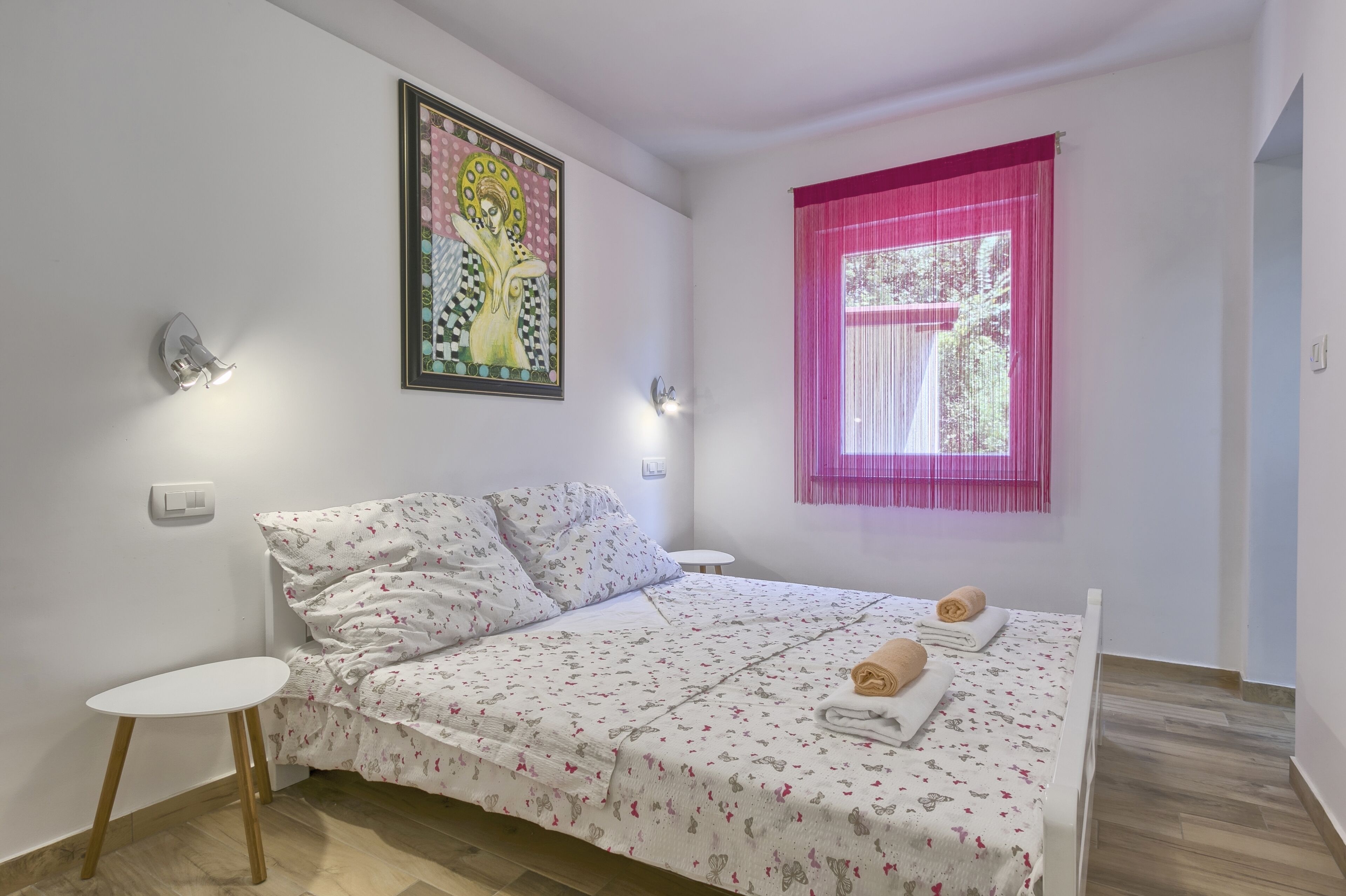 Ein ordentlich bezogenes Bett mit floralem Bettzeug, ein kleiner Nachttisch und eine Wandleuchte.