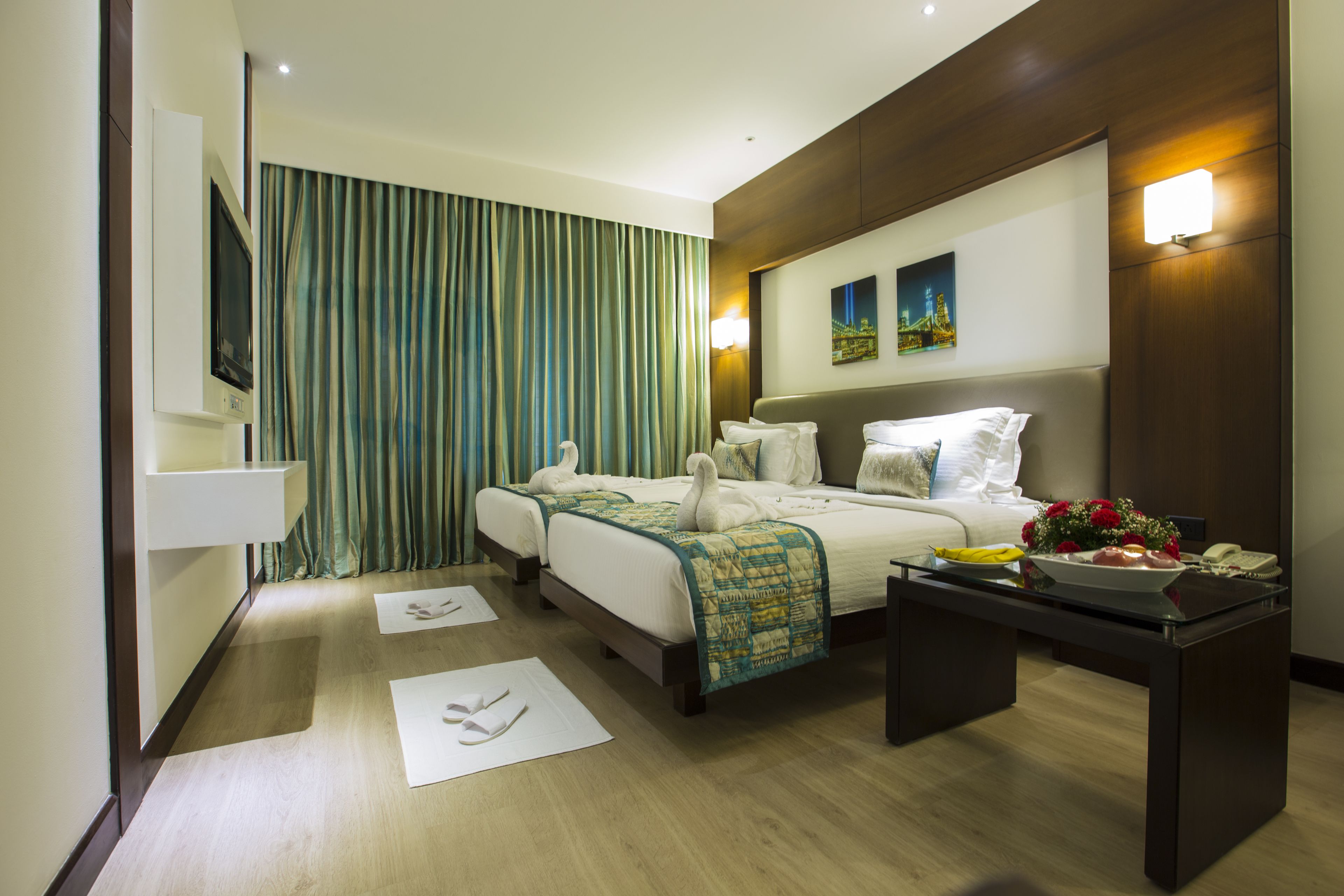 A hotel room with two beds, a flat-screen TV, a bedside table with a bowl of fruit and flowers, and a painting on the wall.