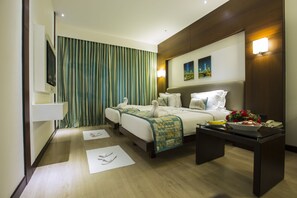 A hotel room with two beds, a flat-screen TV, a bedside table with a bowl of fruit and flowers, and a painting on the wall. - Hash Six Hotels (Coimbatore)