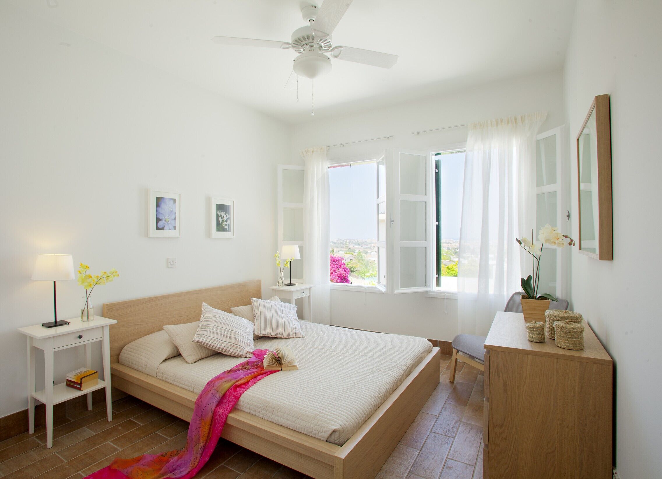 A bedroom with a bed, bedside tables, a ceiling fan, a window with curtains, and a view of the outside.