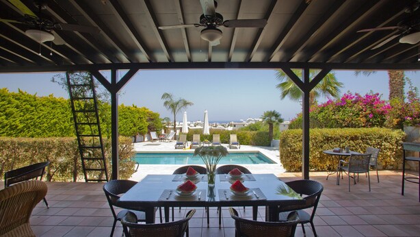 A covered patio with a table set for two, overlooking a swimming pool and a view of the city. - Protaras Pearl With Panoramic Sea View (Protaras)