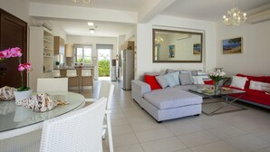 A modern living room with a grey sofa, red and white cushions, a glass coffee table, and a dining area with white chairs and a glass table. - Protaras Villa Posidonas 3 Near The Beach (Protaras)