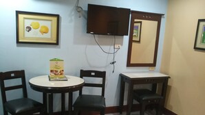 Habitación de hotel con cama, escritorio, dos sillas, una pequeña mesa con un menú, televisor montado en la pared y un cuadro enmarcado.