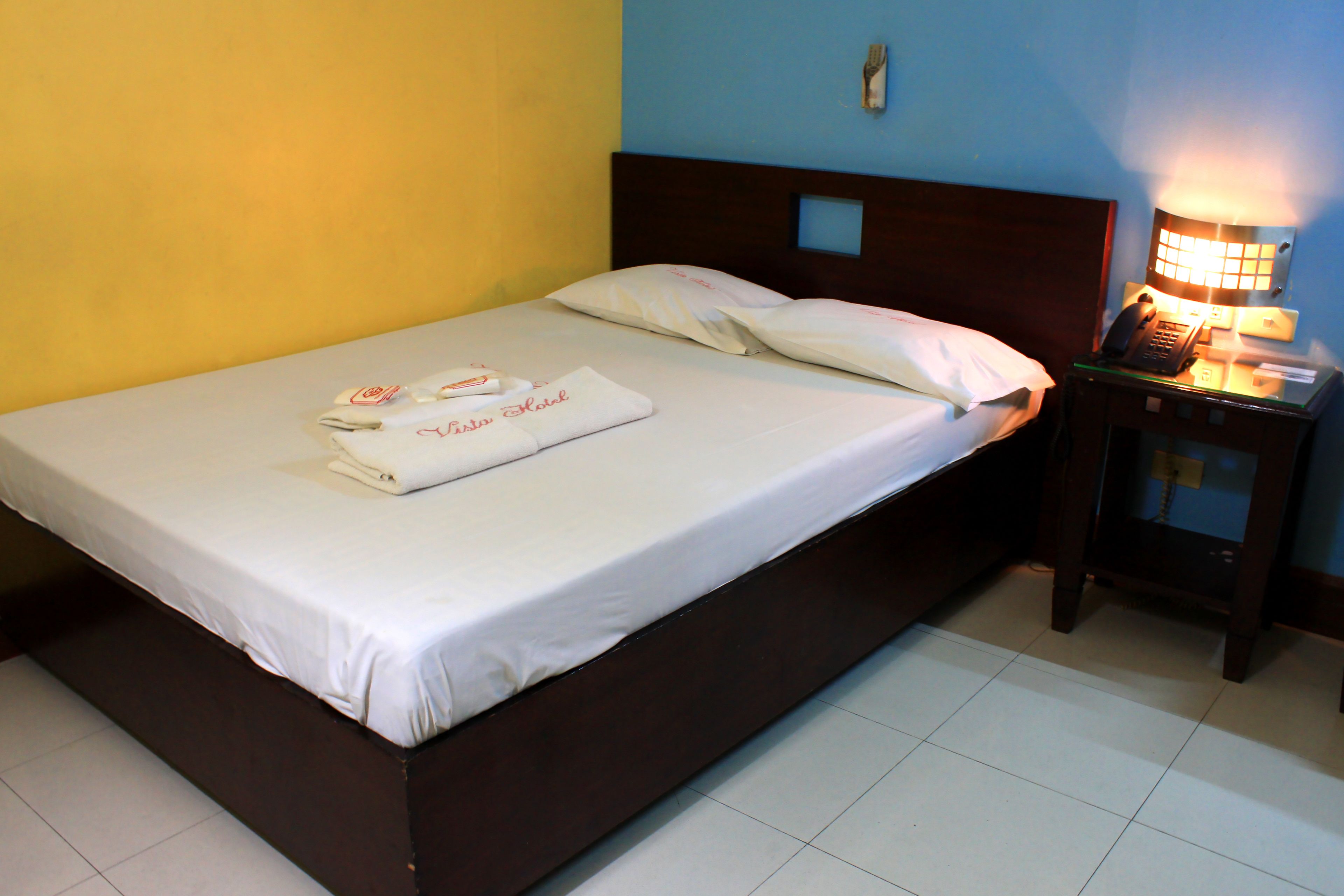 A neatly made bed with white linens and a folded towel, a bedside table with a lamp, and a wall-mounted light switch.