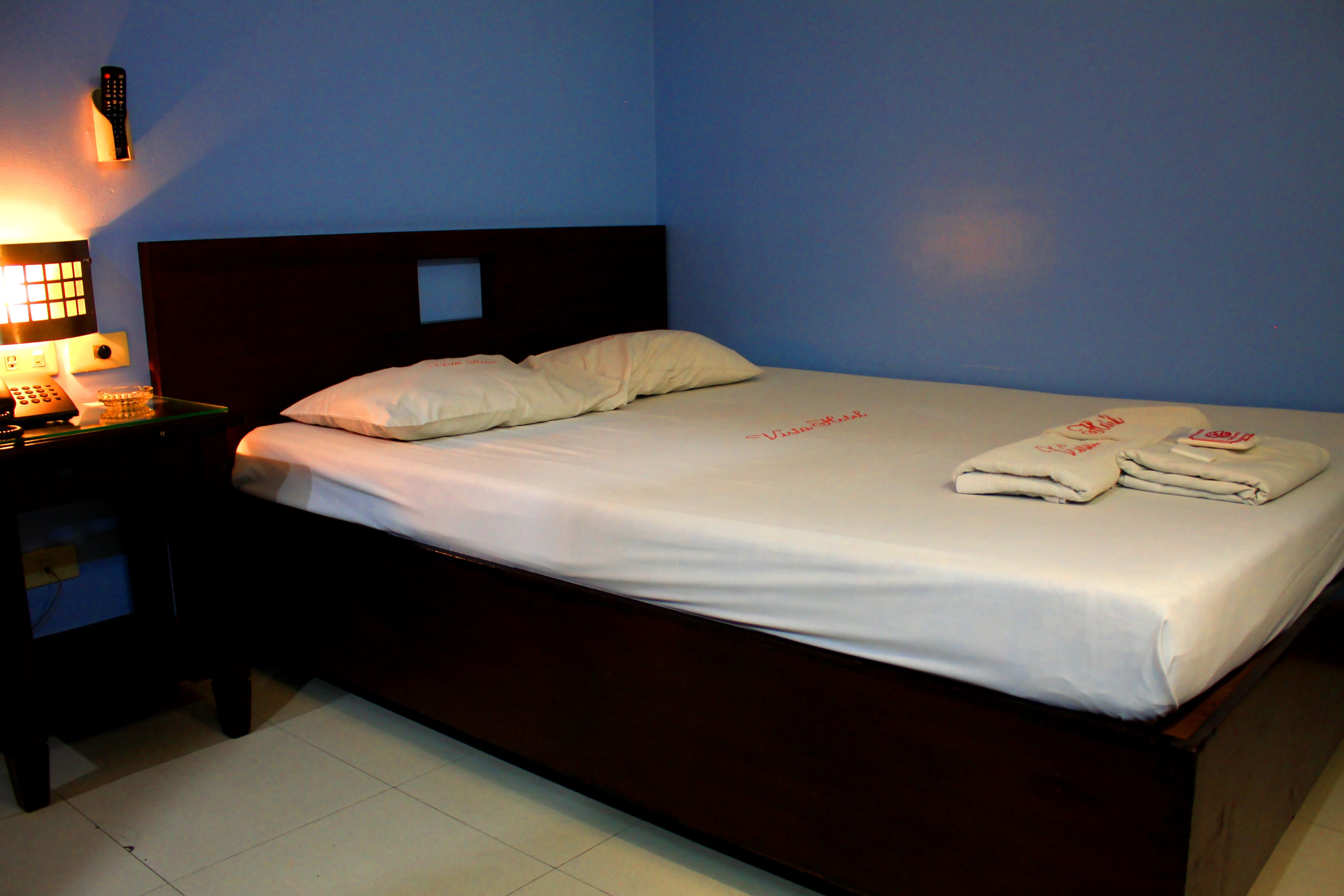 A neatly made bed with white linens and pillows, a bedside table with a lamp, and a remote control on the wall.