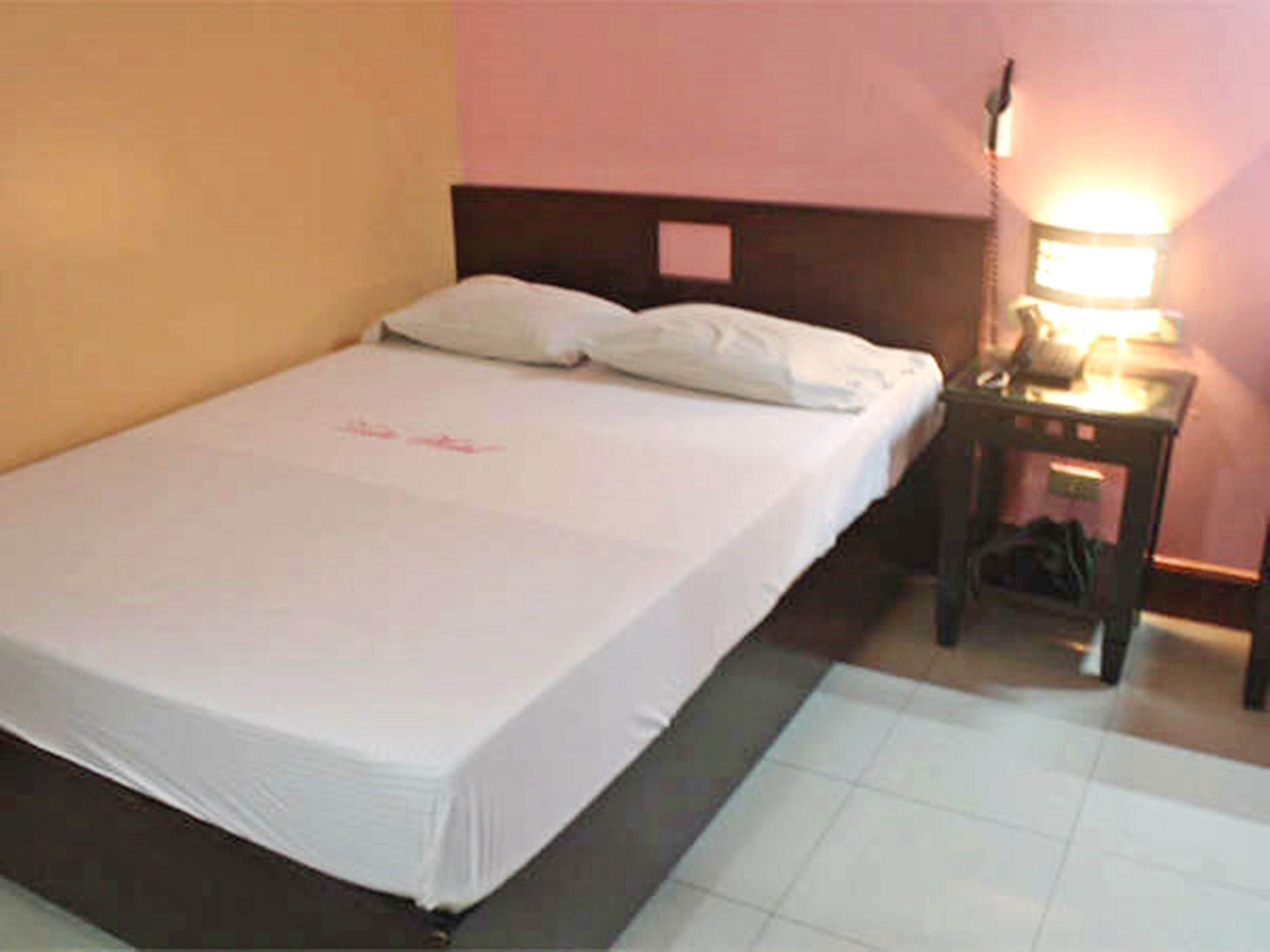 A single bed with white bedding, a nightstand with a lamp, and a tiled floor.