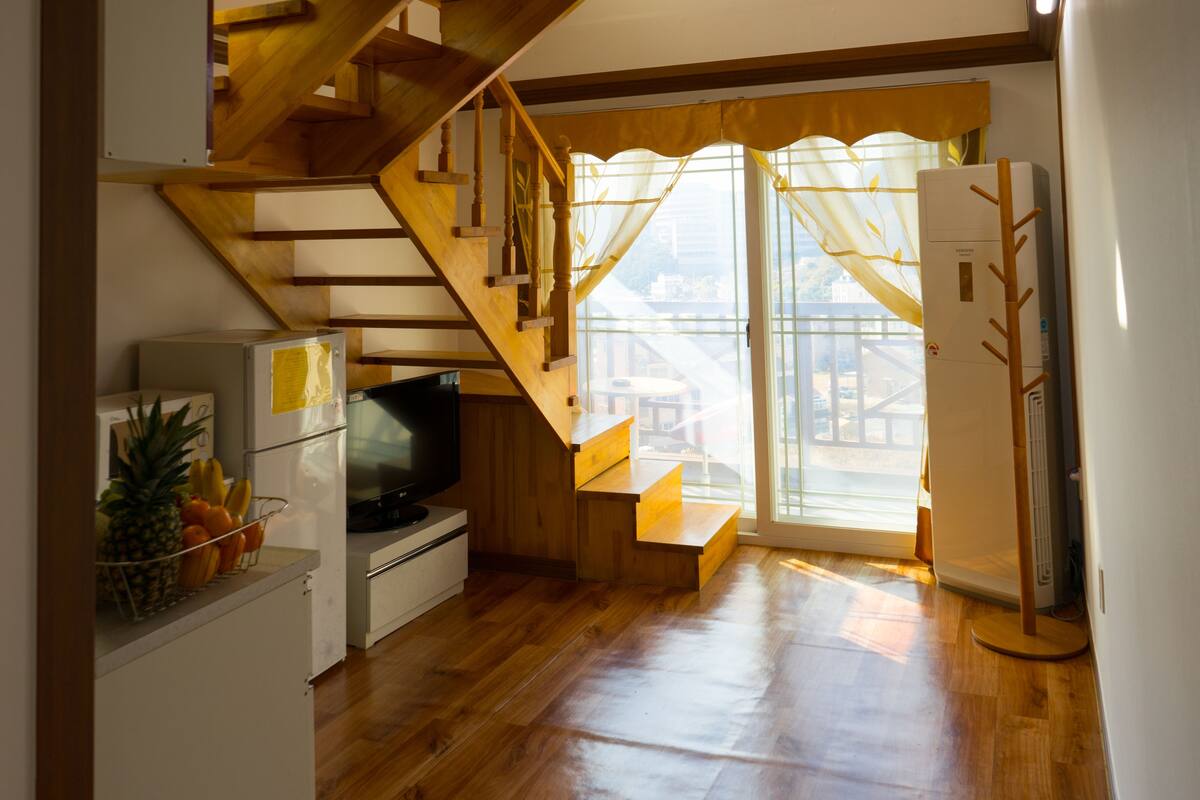 A compact kitchen with wooden flooring, a built-in refrigerator, a small dining area, and a staircase leading to an upper level.
