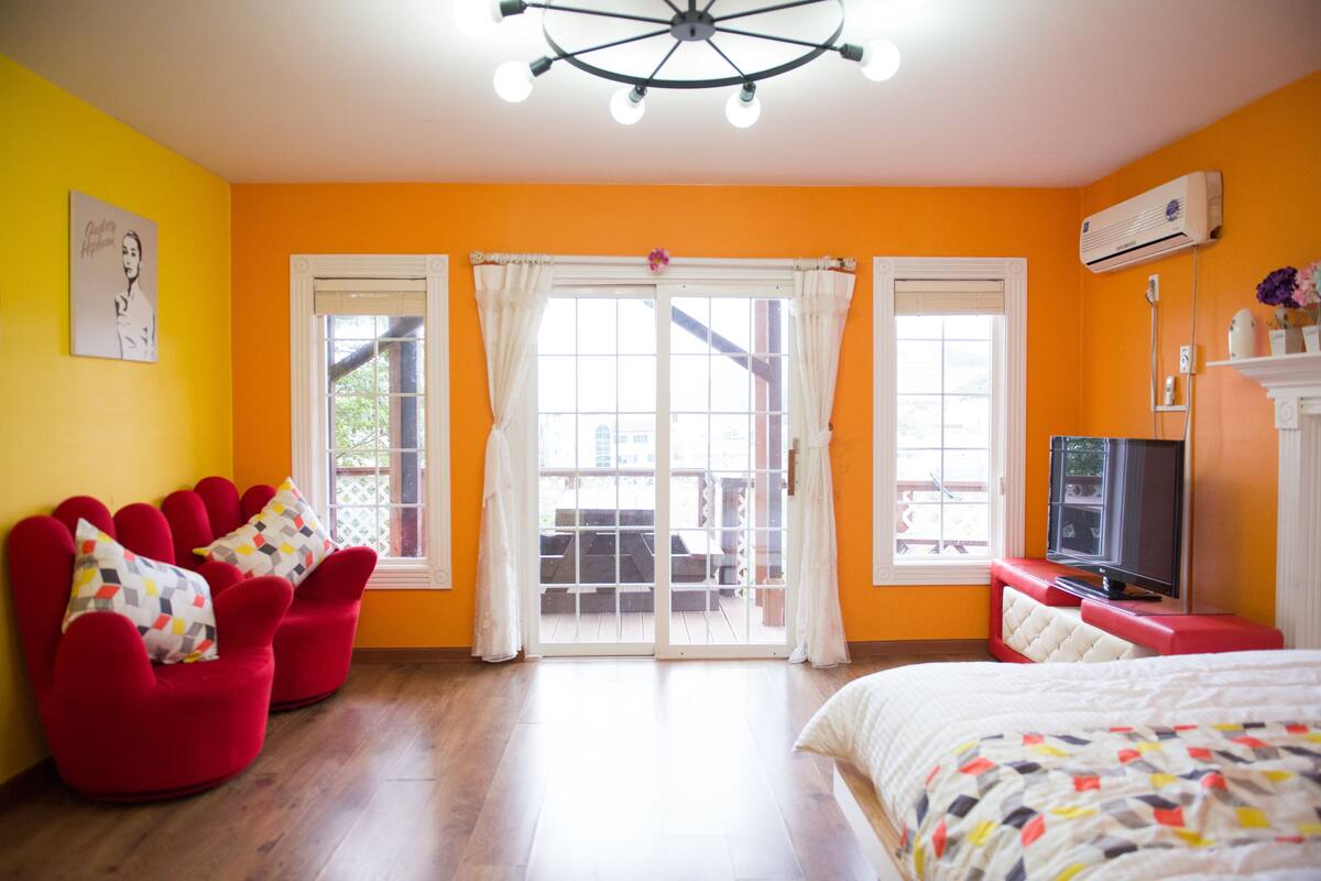 A bright room with orange walls, a bed, a TV, and a sliding glass door leading to an outdoor area.