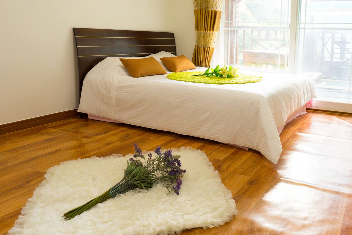 A bedroom with a wooden floor, a bed with white bedding, a brown headboard, and a window with curtains.