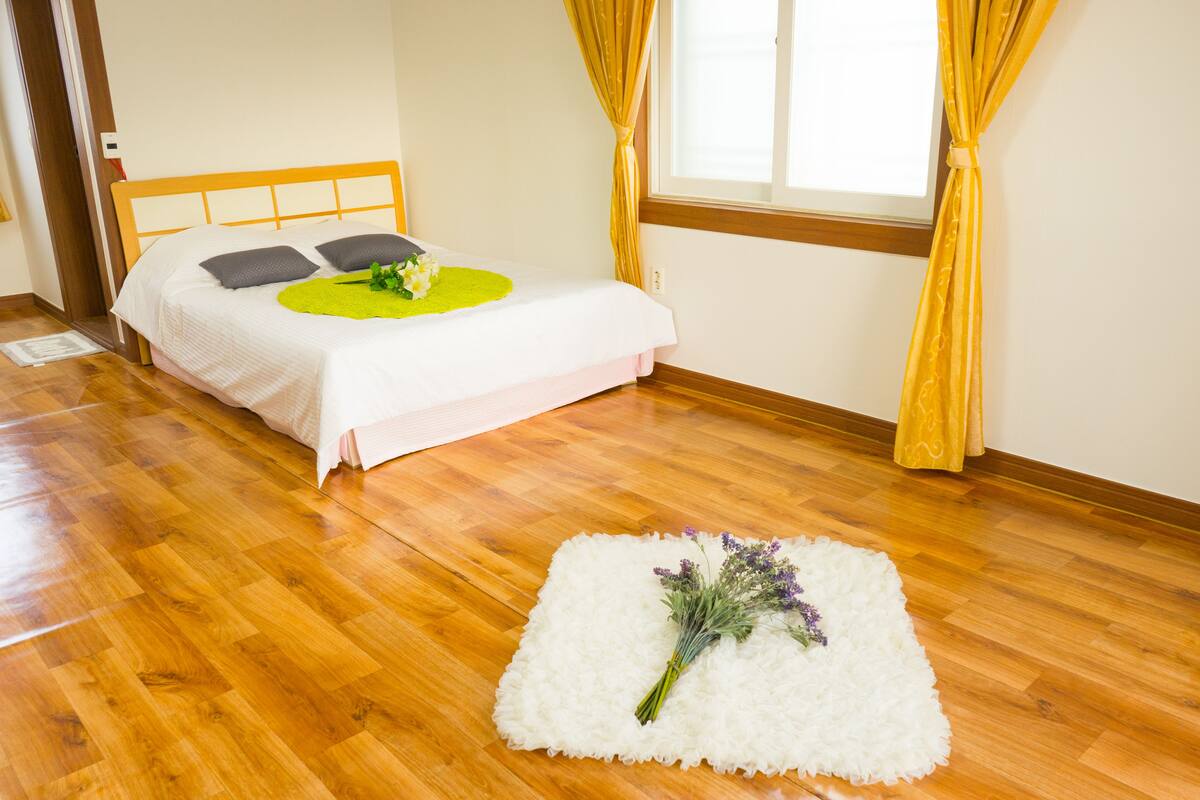 A neatly arranged bedroom with a wooden floor, a bed with a white bedspread, a green circular rug, and a small white rug with flowers.