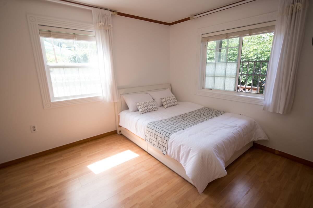 A bedroom with a wooden floor, a white bed with patterned bedding, and two windows with white curtains.