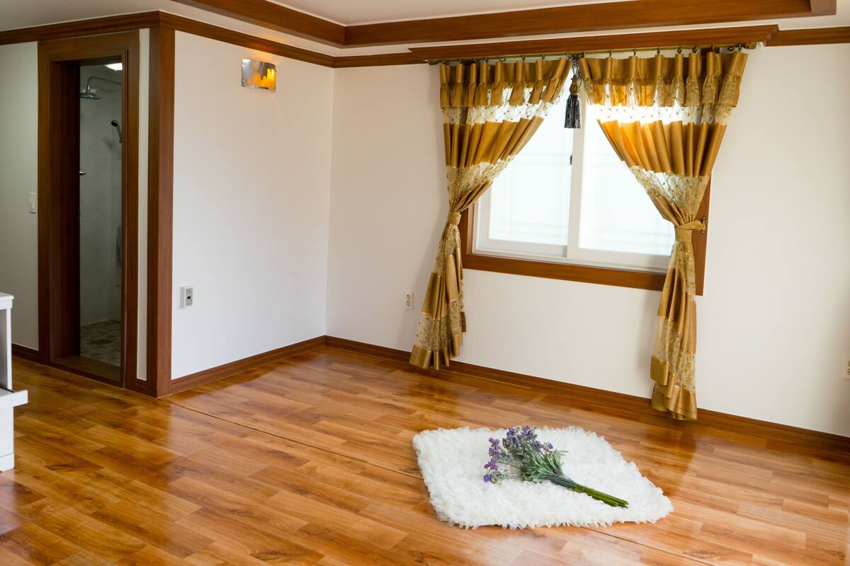 A room with wooden flooring, a window with curtains, and a white rug with a bouquet of flowers.