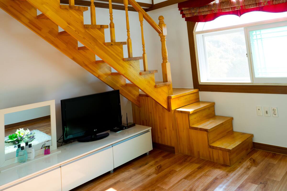 A wooden staircase in a room with a television on a white cabinet, a window with curtains, and a small plant on the cabinet.