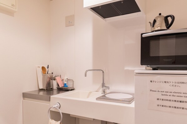 A modern kitchen with a white sink, stainless steel faucet, and a microwave oven. There is a kettle on top of the microwave, and a small shelf with kitchen utensils and a soap dispenser. - COCOSHUKU Tokyo Nakano (Tokyo)